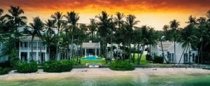 A beachfront mansion with white buildings surrounded by tall palm trees, a central pool area, and a vibrant sunset in the background.