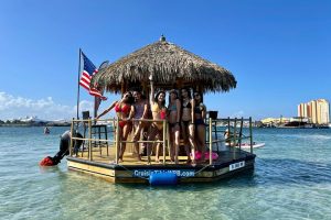 Group of people enjoying time on a cruisin tiki boat with a thatched roof, with buildings on the coastline in the background.