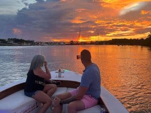 A man and woman sit on a boat at sunset, looking at the view. The man is taking a photo with his phone. The sky is vibrant with shades of orange and yellow, reflecting on the calm water.