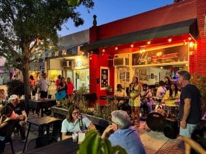 People enjoy outdoor dining and live music outside a red restaurant in the evening, with a band playing and audience seated at tables.