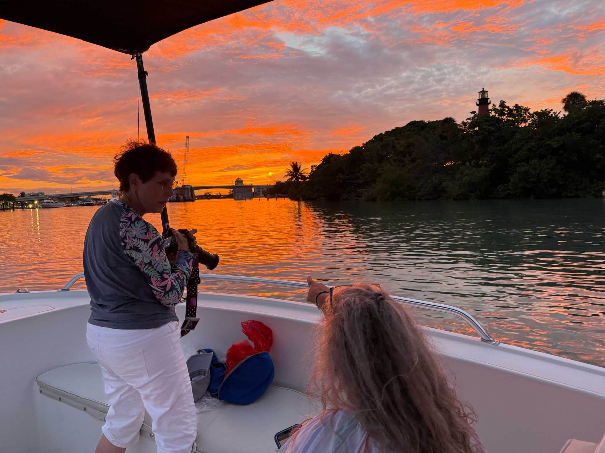 Two women enjoy a sunset on a boat—one holds a camera while the other points toward the vibrant, orange-pink water. Discover scenes like this with West Palm Beach Boat Tours.