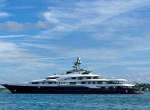 A large, modern white and navy yacht from Palm Beach Mega Yachts is anchored on calm blue water under a partly cloudy sky.