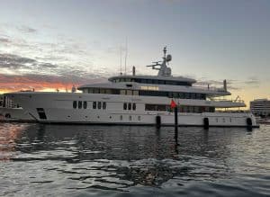 A large white yacht named "TOP FIVE II," one of the stunning Palm Beach Mega Yachts, is docked in a marina at sunset, with calm water and modern buildings in the background.