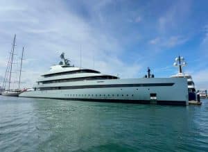 A large modern luxury yacht from Palm Beach Mega Yachts is docked in a marina, with calm water and a clear sky in the background.