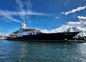 A large, black luxury yacht is docked on calm water under a blue sky with scattered clouds, showcasing the elegance of Palm Beach Mega Yachts.