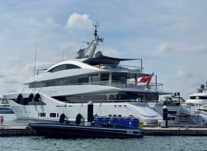 A large white luxury yacht from Palm Beach Mega Yachts is docked at a marina with a red flag at the stern and a smaller boat moored alongside.