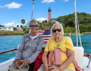 Two women sit on a boat in front of an American flag, with a lighthouse and greenery in the background under a blue sky, enjoying the luxury of Palm Beach Mega Yachts.
