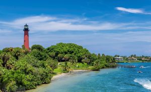 A red lighthouse stands amid trees on the shoreline, as boats and jet skis—including Jupiter boat tours—move across the blue water under a clear sky.